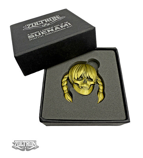 A gold-colored ZOLTRON Sue Nami enamel skull sigil pin with braided hair rests in black foam inside an open black box labeled “ZOLTRON,” with the ZOLTRON art logo also shown in the bottom left corner.