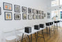 The gallery showcases Cinta Vidals framed Neighbors acrylic on white walls, complemented by Moleskine exhibit catalogs neatly stacked on white folding chairs and tables. Hardwood floors and large windows enhance the space with natural light.