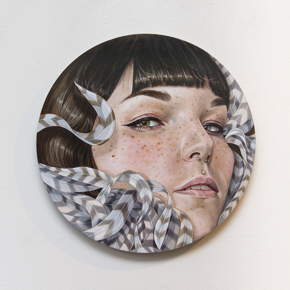 Edith Lebeaus round acrylic on wood painting showcases a close-up of a person with short dark hair, freckles, and a nose ring. Curved, ribbon-like geometric patterns wrap around the face like a feather cocoon on a plain white background.
