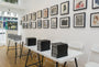 The gallery interior features Zachary Oldenkamp custom-framed artworks on the walls. In the center, two tables hold stacks of black books, emulating a Moleskine exhibit, surrounded by white folding chairs. A window grants a view of the street outside.