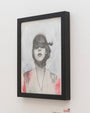 A framed monochrome portrait by Nimit Malavia features a subject with hair covering their eyes against a light background enhanced by subtle pink and gray brushstrokes. The simple black frame complements the artworks enchanting Nightwalks vibe.