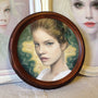 A framed portrait by Bec Winnel features a woman with brown hair and a serious expression against a green and gold backdrop. She wears a white top, surrounded by a round wooden frame hinting at two partially visible paintings, creating an ambiance reminiscent of Spoke Art Gallery.