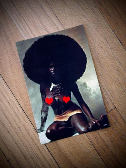A photograph of a person with a large afro kneels on a wooden floor, wearing red heart shapes on their chest and yellow shorts. Holding a cigarette with visible smoke, the scene evokes Shining-inspired mini prints from Tim Doyle, adding an intriguing layer.