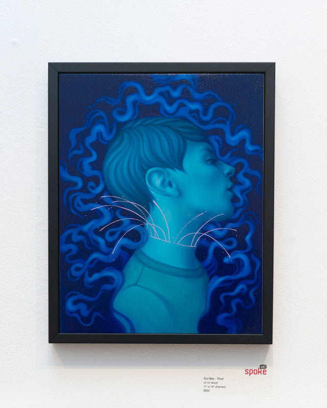 Surreal blue-themed Syd Bee painting features a profile with closed eyes, encircled by wavy tendrils and intersecting white neck lines. Displayed at the Spoke Art gallery, the artwork is elegantly framed in black against a white wall.