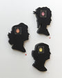 Three intricately crafted black silhouette wall hangings of womens profiles by Matt Ritchie, made from hand-cut wood featuring a small embroidered portrait at face height, are mounted on a light-colored wall like an art gallery display with small red labels beneath them.
