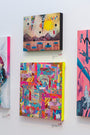 A colorful art gallery wall by Haunted Euth features three abstract paintings. The top piece displays warm-toned geometric shapes, the middle is alive with vibrant patchwork patterns, and the right painting blends pink and blue in a captivating abstract dance.