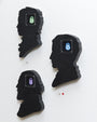 Three Matt Ritchie black silhouette wall sculptures with colorful robot faces in green, purple, and blue are mounted on a white wall. Each hand-cut wood piece features a unique profile, offering a modern and playful touch.