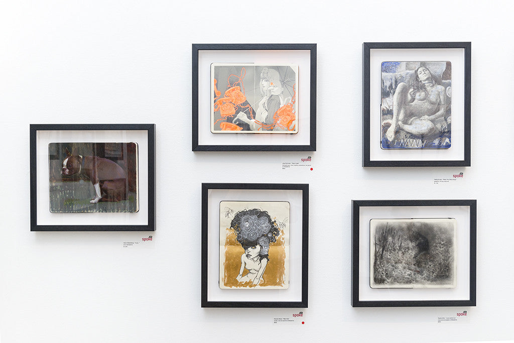 A display of five framed Randy Ortiz artworks, akin to a Moleskine exhibit, adorns a white wall. These pieces blend abstract and representational styles with diverse colors and textures, featuring figures and organic forms that evoke the haunting allure of Lost Lambs Cry.