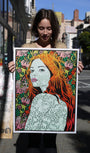 On a city street, a woman holds a vibrant Chuck Sperry print featuring a red-haired woman with floral surroundings. This limited edition artwork highlights intricate facial and clothing patterns, embodying modern urban artistry.