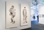 Two large, detailed monochrome portraits by Joel Phillips in charcoal and graphite hang on a white gallery wall. The left shows a rugged figure in winter clothing, the right depicts someone in casual wear. A visitor views other artworks reminiscent of San Francisco art fairs in the background.