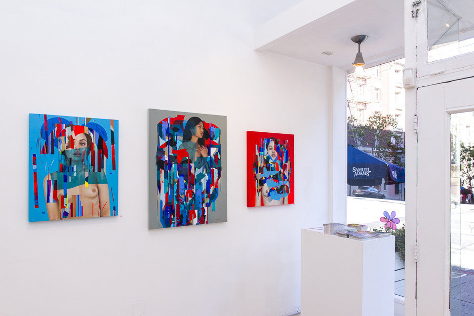 Three vibrant abstract paintings by Erik Jones featuring partly hidden figures adorn a white gallery wall, with natural light highlighting a small pedestal displaying brochures and a decorative flower.