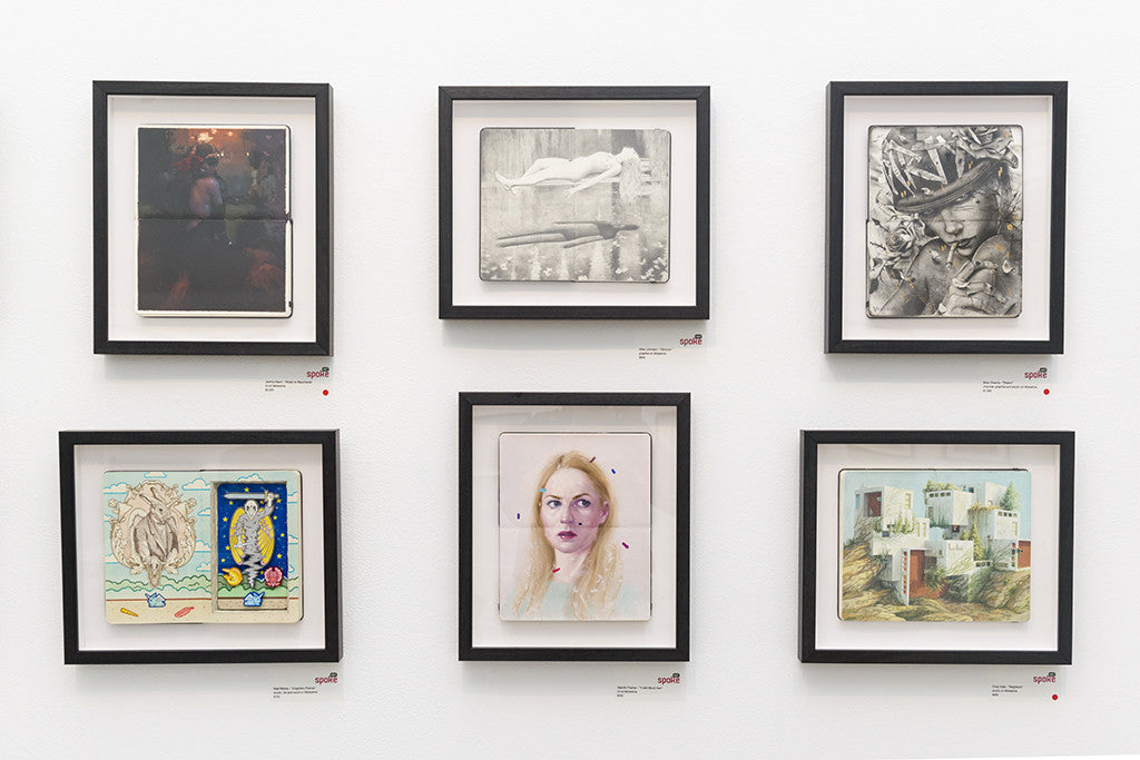 A gallery wall features six framed artworks from Brian M. Viveros, showcasing diverse styles such as abstract, portrait, and landscape. Each uniquely colored piece in simple black frames against a white wall subtly echoes the evocative essence of Disarm.