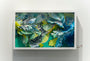 A Crystal Wagner-inspired shadow box art piece showcases intricate paper sculptures with vibrant green, blue, and yellow layers reminiscent of abstract foliage in Terrarium II. The boxs shadows add depth to the swirling patterns.