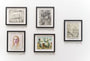 The gallery wall, featuring Cinta Vidals five framed artworks with a mix of black-and-white sketches and vibrant colored portraits arranged asymmetrically, is accompanied by small labels with red dots. The white wall beautifully enhances this Moleskine exhibits visibility.