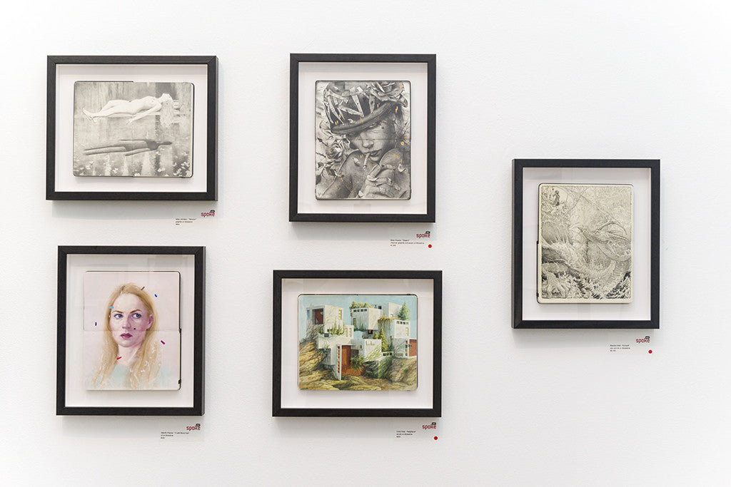The gallery wall, featuring Cinta Vidals five framed artworks with a mix of black-and-white sketches and vibrant colored portraits arranged asymmetrically, is accompanied by small labels with red dots. The white wall beautifully enhances this Moleskine exhibits visibility.