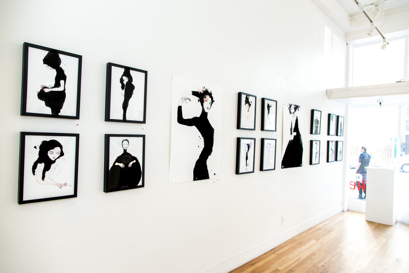 A gallery wall displays monochrome paintings inspired by Conrad Roset, featuring abstract silhouetted figures. Created with India ink, these artworks enhance a wooden-floored room with white walls and natural light streaming through a glass door on the right.