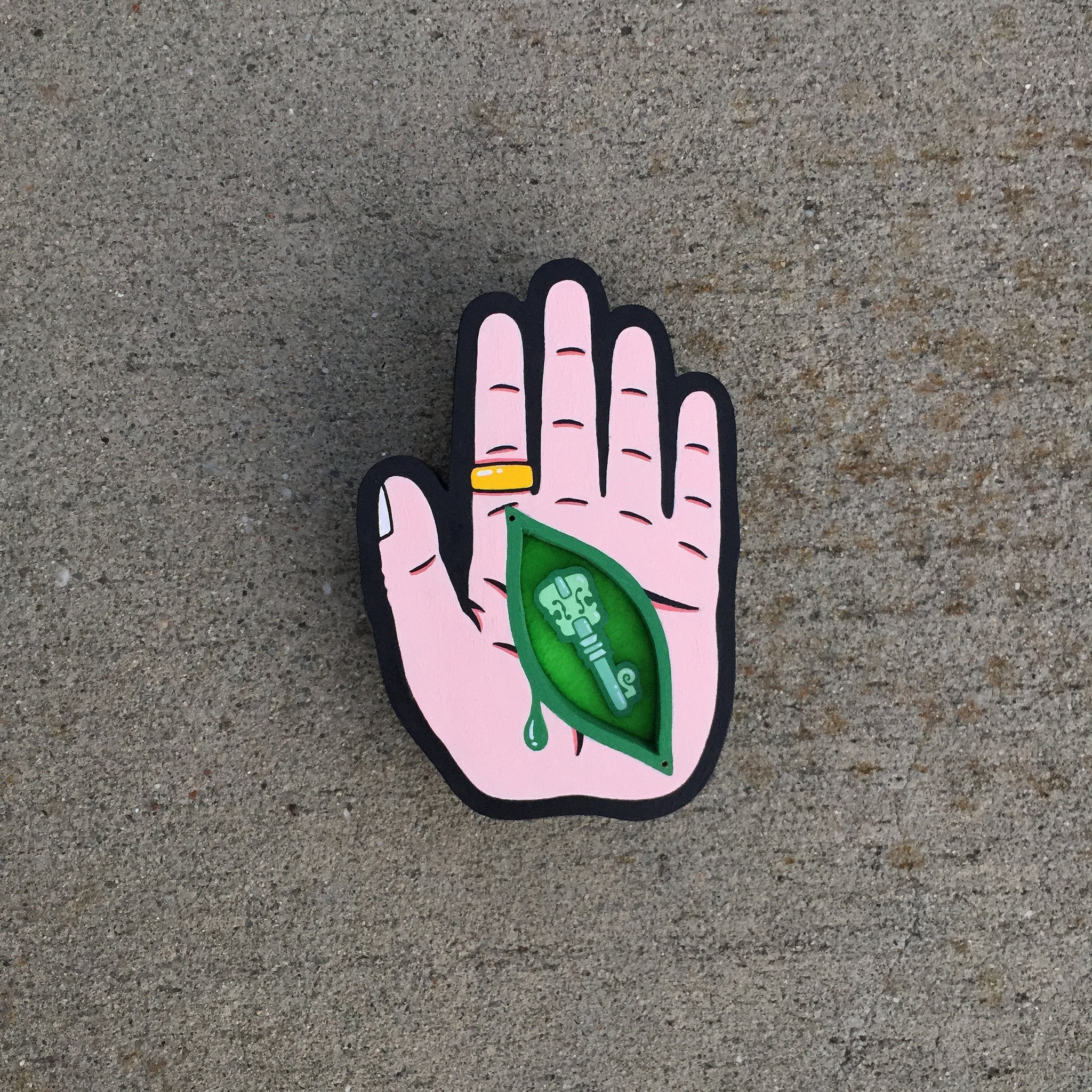 Titled Idol Fun, this whimsical Matt Ritchie illustration features a pink hand with a gold ring and white fingernail. A green, leaf-shaped eye design with a drop beneath it adorns the palm. Set against textured gray concrete, it uses acrylic on cut wood for depth and charm.