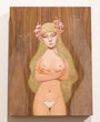A serene woman with long green hair, adorned with pink flowers, stands topless with arms crossed in white underwear against a wood-textured background. This soft, ethereal oil on wood piece by UYU is reminiscent of art showcased at Spoke Art Gallery.