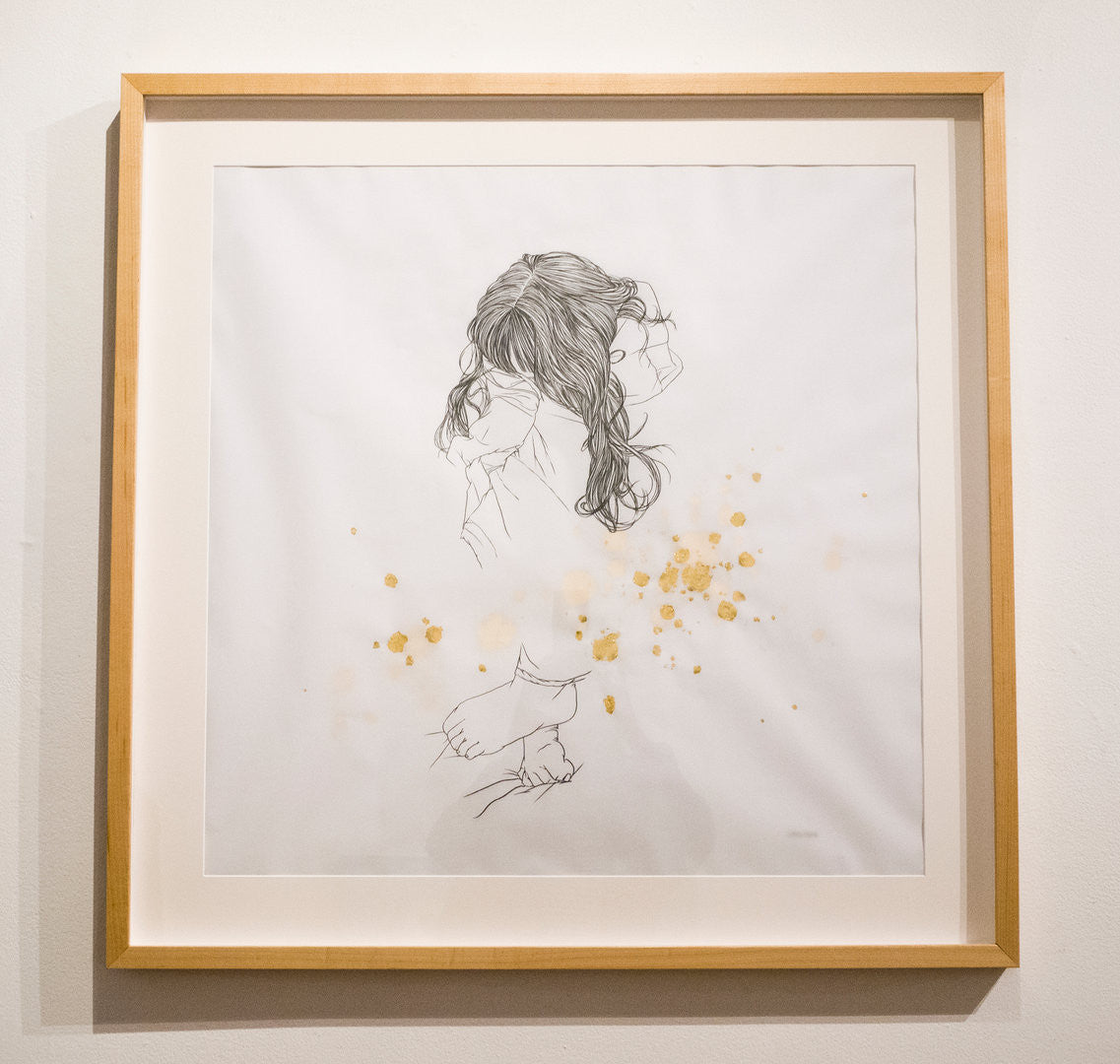 Christine Wus framed artwork presents a minimalist sketch of a woman with long hair, her face partly turned away. Pencil and gold leaf splatters add vibrant color, enhancing the monochrome drawing with gentle flair.