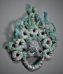 The Silver Streaks Mask by Lucien Shapiro is a decorative piece with a textured, scale-like surface, intricate spirals, and greenish-blue crystals. It features eye holes and is set against a dark background similar to street diamonds.