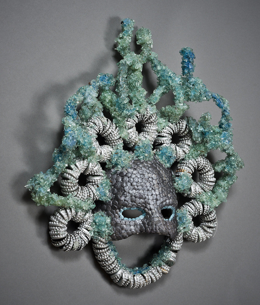 The Silver Streaks Mask by Lucien Shapiro is a decorative piece with a textured, scale-like surface, intricate spirals, and greenish-blue crystals. It features eye holes and is set against a dark background similar to street diamonds.