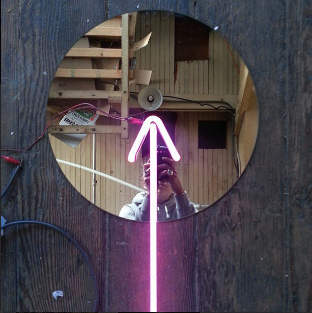 A circular mirror on a wooden floor reflects someone capturing the moment, embodying the spirit of a seeker of inner truth. A neon pink arrow, reminiscent of Meryl Patakys art, points upwards against a backdrop of vintage wood, speaker, and staircase.