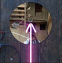 A circular mirror on a wooden floor reflects someone capturing the moment, embodying the spirit of a seeker of inner truth. A neon pink arrow, reminiscent of Meryl Patakys art, points upwards against a backdrop of vintage wood, speaker, and staircase.