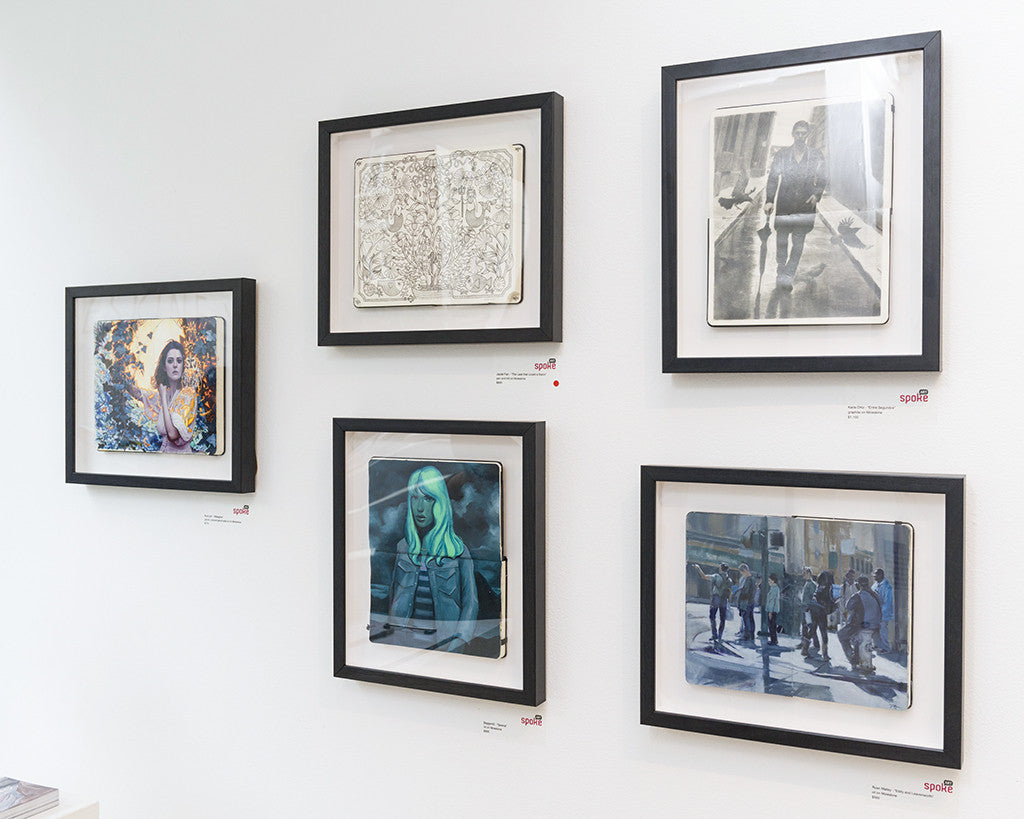 The gallery wall, featuring the Ryan Malley collection, showcases five framed artworks including portraits, a sketch, and a street scene. Each piece is individually framed and displayed on a white wall with small labels beside them.