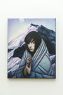 This Michael Shapcott oil on linen painting portrays a tranquil figure in a flowing gray cloak, standing serenely before snowy mountains beneath a pale sky. Dark hair frames their face as moonlight casts its glow—a true Hyperballad of peace.