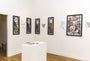 An art gallery room presents Rhys Coopers screen prints of women on white walls. In the center, a white pedestal holds open books, possibly Alice tales, while a wooden floor guides visitors to another inviting room.