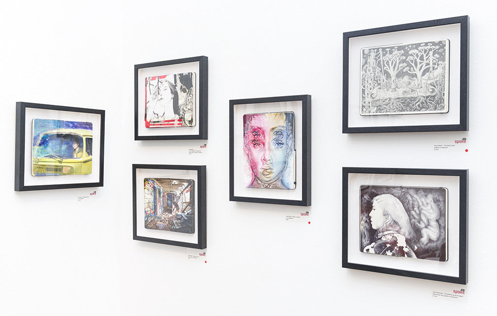 A gallery wall at Spoke Art presents six framed artworks by Ben Smith, featuring vibrant portraits and abstract scenes. Uniformly spaced black frames create a balanced display against the white wall, with small labels offering insights into these captivating pieces.
