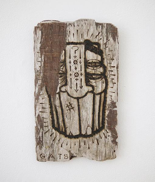 A rustic wooden artwork by GATS, carved from burnt driftwood featuring a face with eyes and symbolic patterns such as arrows and a question mark. It measures 6.25 x 10.5 and is signed GATS at the bottom, showcasing a textured, aged appearance.