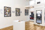 Inside a small art gallery, Rhys Coopers framed illustrations adorn white walls, featuring intricate screen prints of women and ravens. A central pedestal displays more art pieces. A glass door with signage provides access outside, while natural light enhances the shine of the wooden floor.