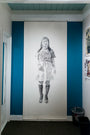 A detailed charcoal and graphite drawing by Joel Phillips of a person in a dress and knee-high socks decorates a blue accent wall. It spans from ceiling to floor, with a small trash can on the right.