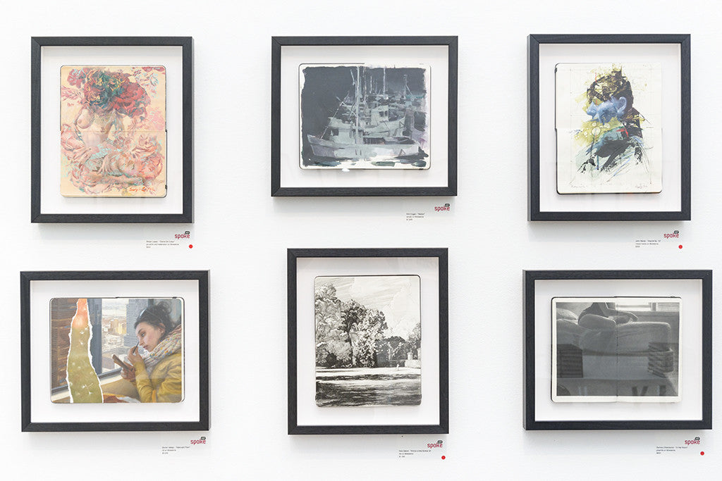 A wall displays six custom framed artworks by Zachary Oldenkamp: top row has abstract and ship paintings; bottom row features a person on phone, a graphite tree scene in black and white, and another abstract. Each piece is labeled with a red dot below.