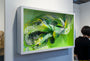 Crystal Wagners framed 3D art, Terrarium I, exhibits an intricate swirling pattern with layered paper cuts in green, yellow, and white. Admired by two gallery visitors at Spoke Art, it captivates with its complexity.