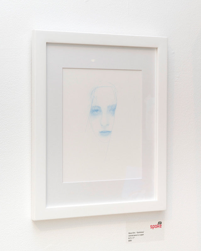 On a white wall, framed artwork by Steve Kim showcases a minimalist face sketch in soft blue tones with light, wispy colored pencil strokes emphasizing the eyes and lips. A label below marks its selection for the Supersonic Invitational.