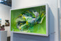 A 3D abstract art piece by Crystal Wagner, similar to her Terrarium I series, is framed with a white border. It showcases intricate layers of swirling shapes in white, yellow, and blue against a vibrant green backdrop and is elegantly displayed at the Spoke Art gallery.