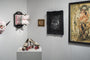 An art gallery display showcases an ornate high-heeled shoe sculpture with pink roller skate wheels on a pedestal, surrounded by framed artworks. Among them is a mixed media piece and intricate wall art in antique frames by Handiedan.