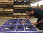 A tattooed person kneels, carefully painting black bear designs on purple posters. Limited edition Eelus art prints are rolled up and stacked behind them as they work with intense focus.
