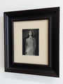 A dark wood-framed black and white portrait of a woman in a dress from the Chimerical exhibition by Karla Ortiz is displayed on a white wall.