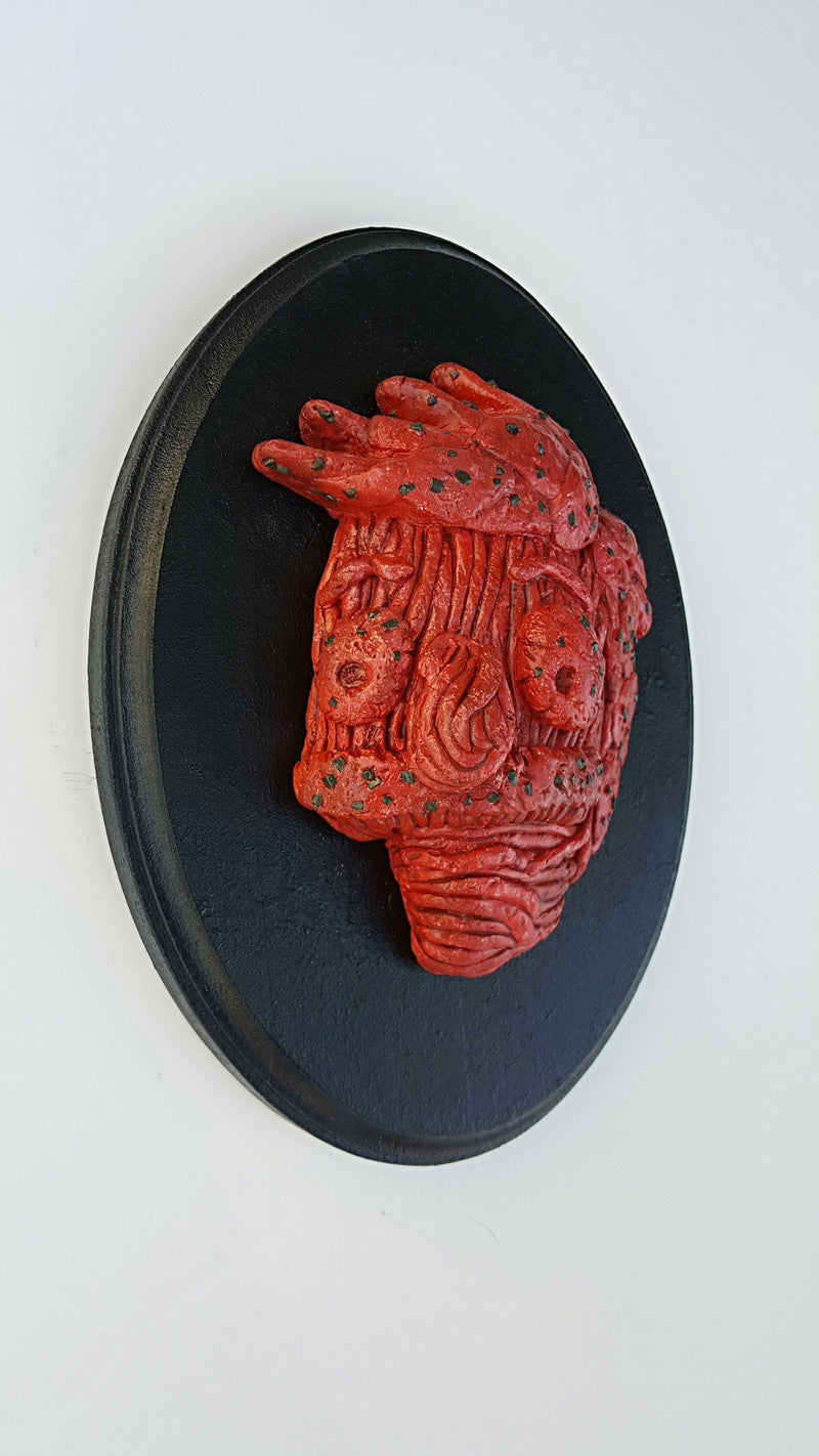 The Meathead by Eric Price is a red, textured clay sculpture featuring intricate grooves and dark speckles. Its mounted on a black circular base against a white wall and evokes the abstract or animalistic faces often seen in Bobs Burgers art shows.