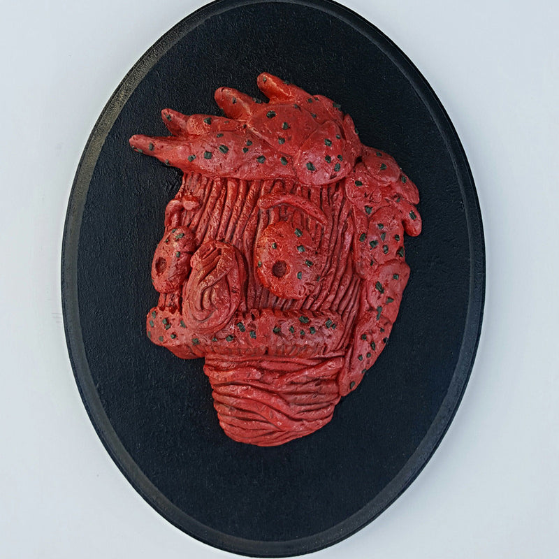A red abstract bust titled Meathead is set on a round black plaque. Its textured, swirling patterns and dark specks create a stylized profile, reminiscent of Eric Prices evocative work displayed at Bobs Burgers art show.
