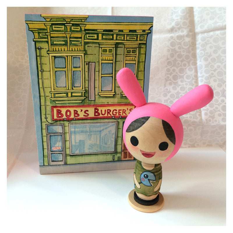 A wooden doll wearing a pink rabbit-ear hat stands in front of a Bobs Burgers themed box, perfectly capturing Alina Chaus whimsical watercolor style. The doll has a cartoonish face and features a small bird design on its outfit.