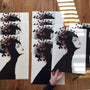 A person holds a Conrad Roset fine art giclée print of a woman with an elaborate floral headdress. The table is lined with more limited edition prints, as sunlight creates depth and contrast through shadows.