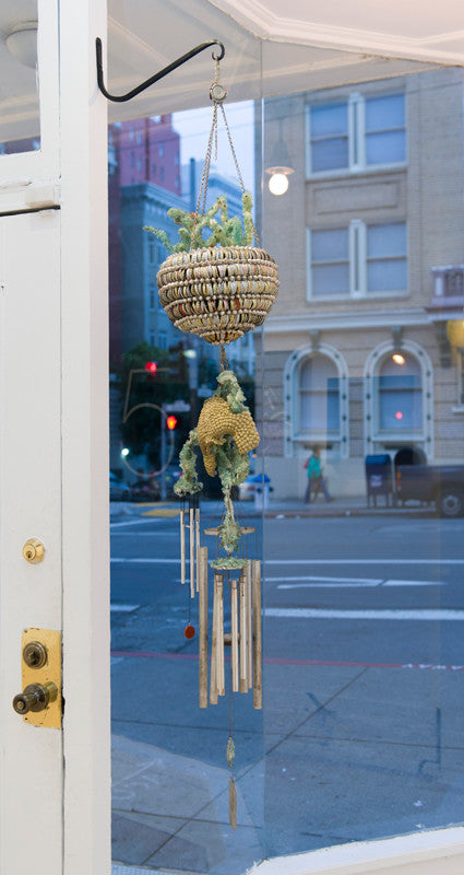 In the doorway, a Lucien Shapiro decorative wind chime with its woven basket and metal tubes sways gently. Through the window, blurred buildings and people blend urban life with delicate artistry, subtly nodding to Shapiros creative touch.