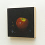 A small square acrylic painting on wood, reminiscent of Lilly Piris style, depicts a glossy red apple against a black background on a light wall. It features delicate details like marbles and a butterfly nearby.