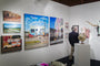 In an art gallery, a person in a dark blazer admires Jessica Hesss vibrant outdoor oil on canvas paintings on white walls, complemented by a globe and an antler sculpture in the foreground.