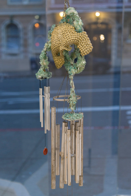 Lucien Shapiros abstract sculpture, with textured yellow and green organic shapes on metal rods and chimes, hangs in front of a window. The background reveals a blurred street scene through the glass, capturing the vibe of the Supersonic Invitational.