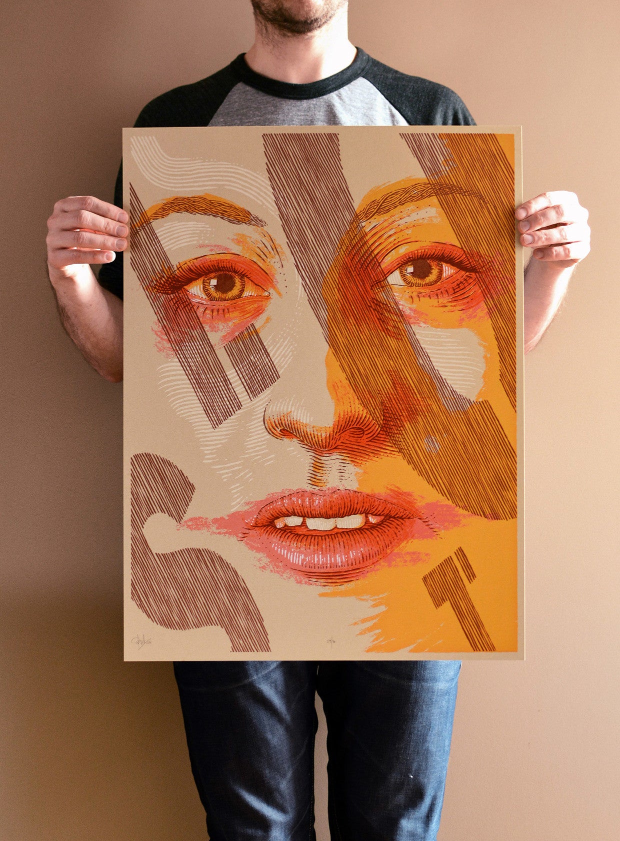 A person holds a Bartosz Kosowski limited edition art print, showcasing a stylized portrait of a womans face with abstract patterns and bold letters. The warm orange and brown background perfectly complements their casual shirt and jeans.
